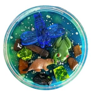 Rond doosje gevuld met groene en blauwe sensorische klei, versierd met een plastic libelle, bever, kikker en kleurrijke steentjes, boomstammen en glitter – een zoemende sloot- of rivierzijde in miniatuurformaat.